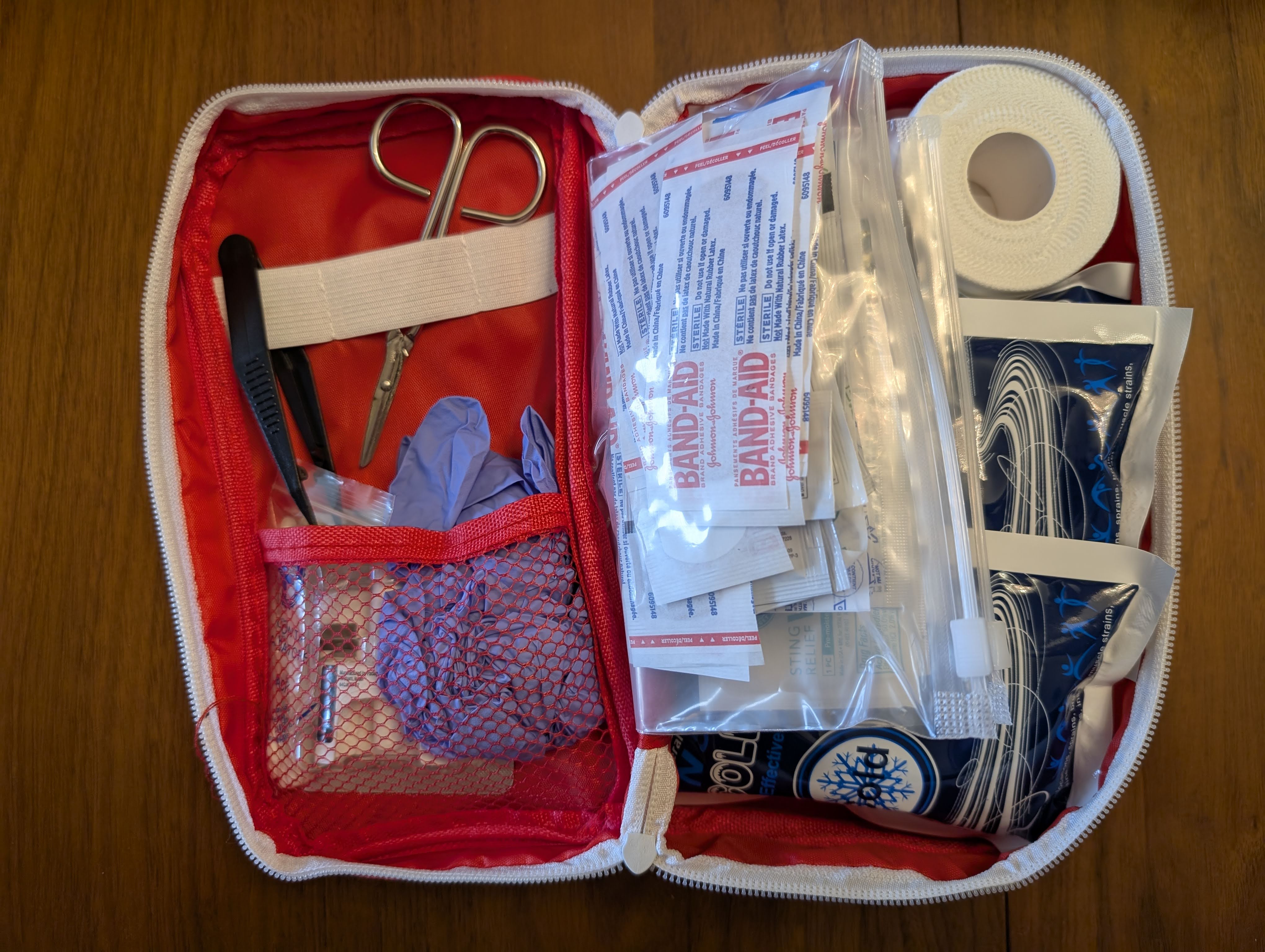 Open first aid kit showing organized supplies including bandages, gloves, scissors, tweezers, gauze pads, tape, and instant cold packs inside a red zippered pouch.