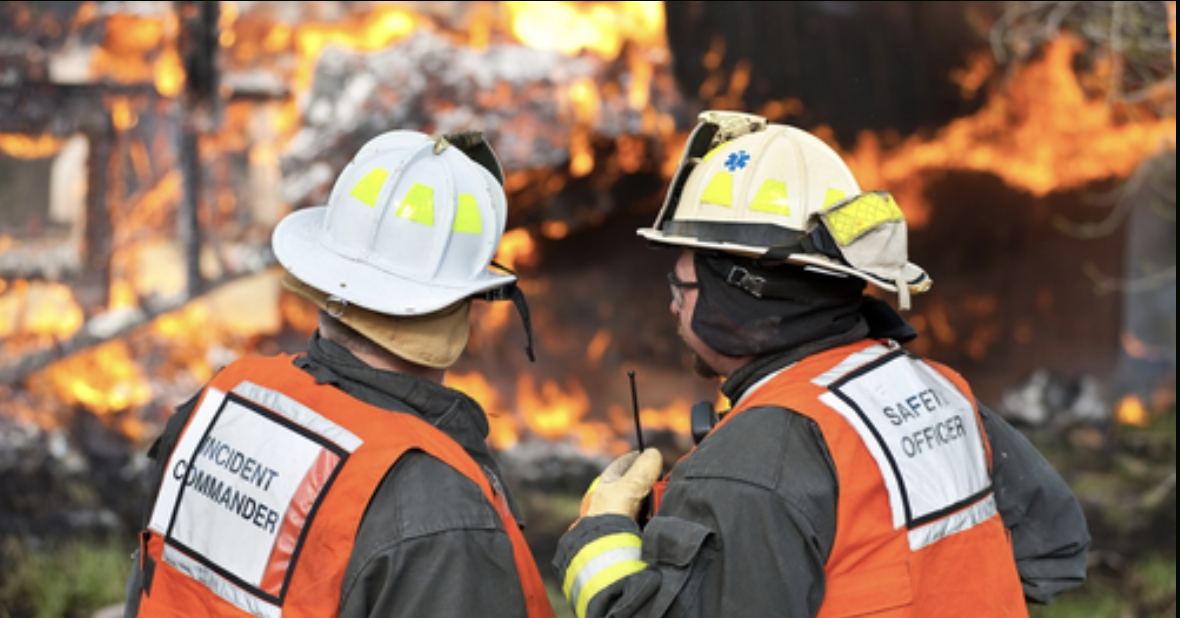 Two firefighters in command vests at an active wildfire scene, one wearing an &ldquo;Incident Commander&rdquo; vest